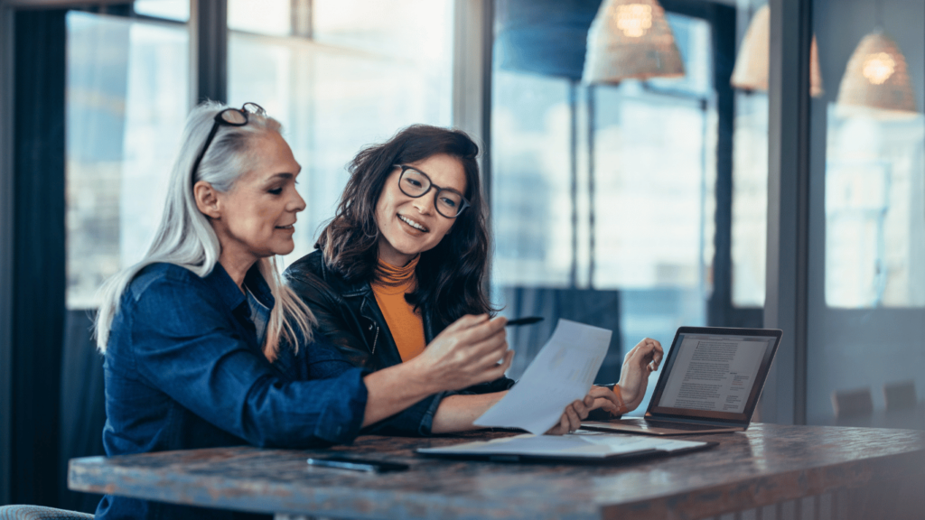 The image shows one woman helping another woman at work. Edgility helps organizations develop and implement executive onboarding plans that keep their mission moving during transitions.