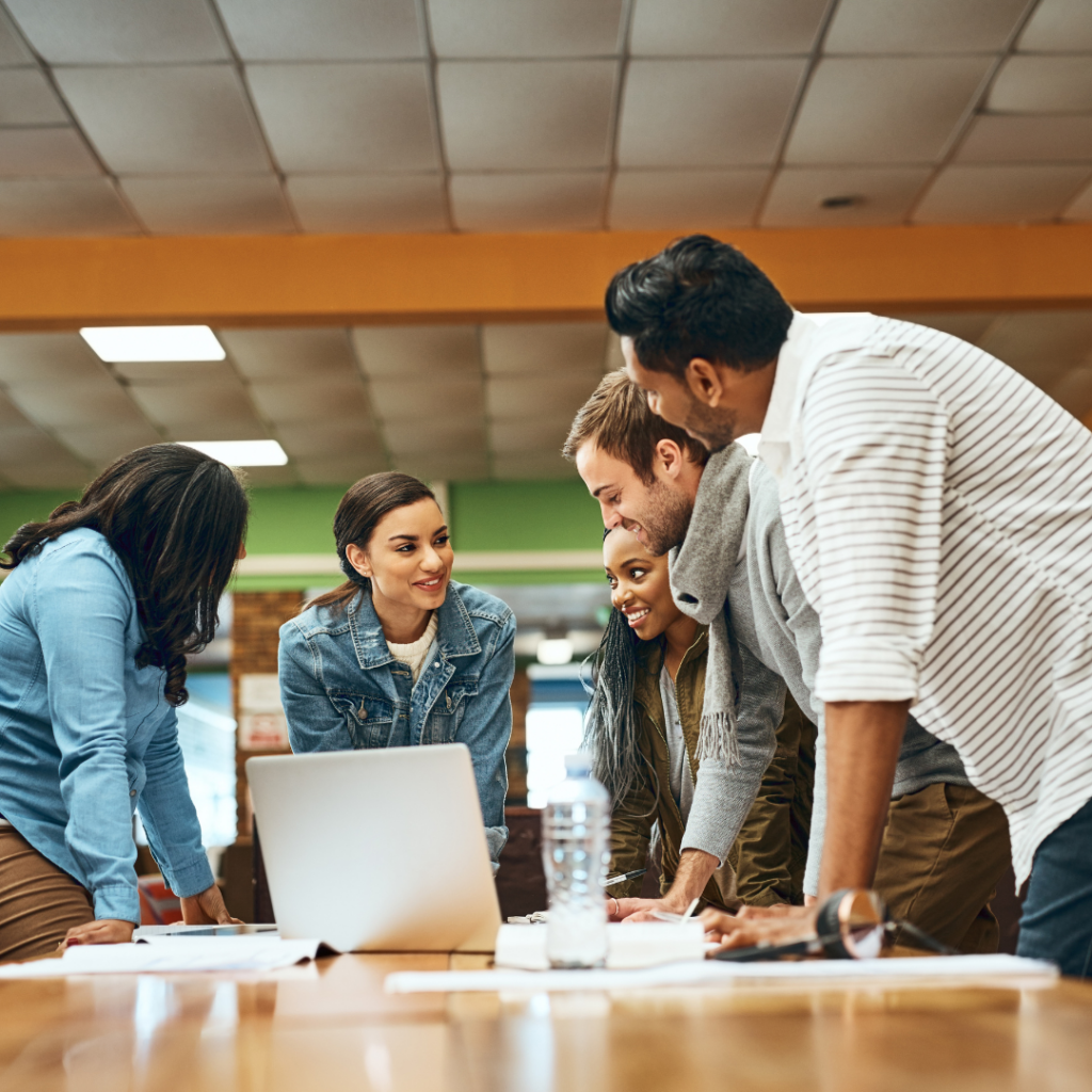 The image shows coworkers gathering around a laptop. Edgility offers compensation design for nonprofits, schools and small businesses